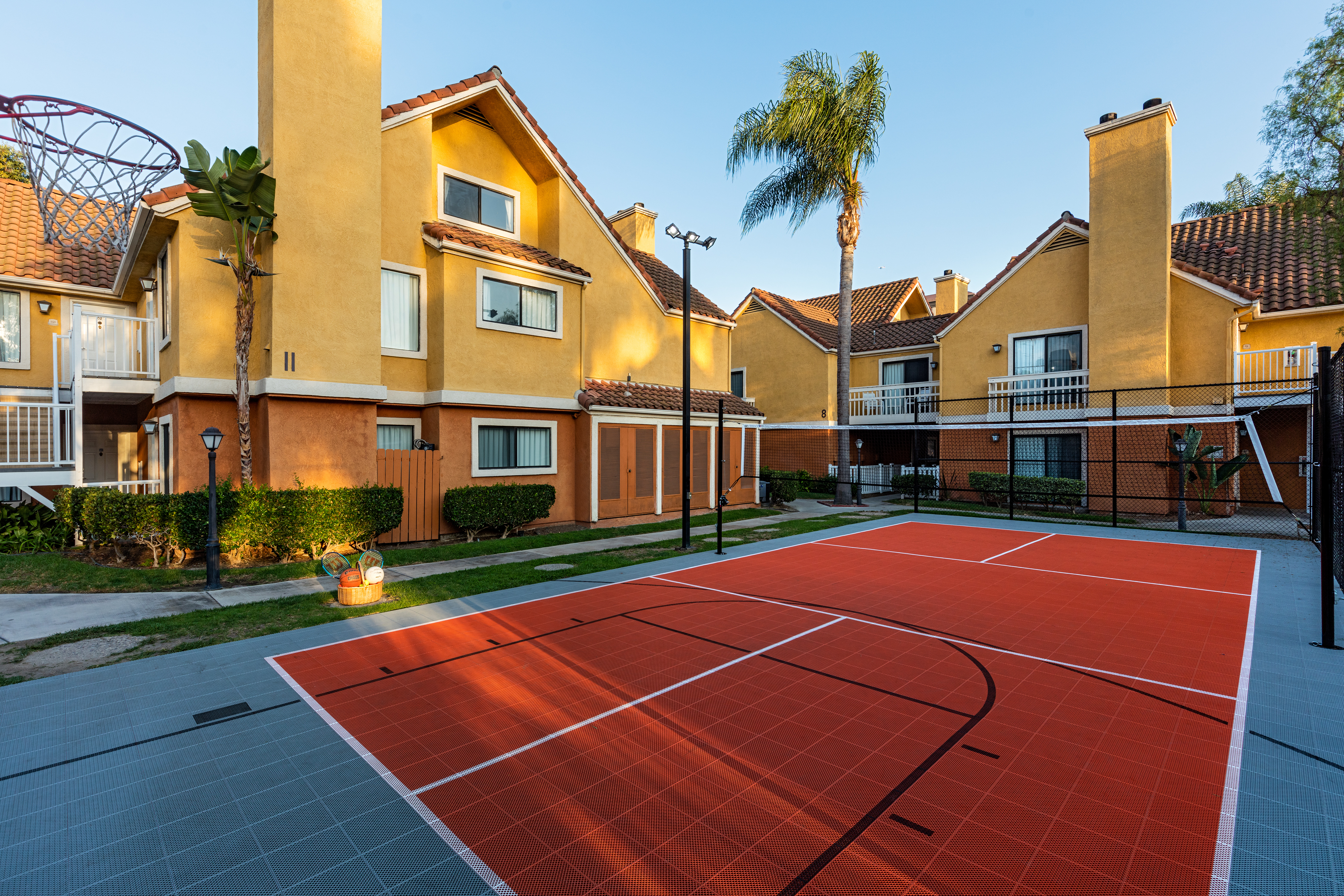 A red basketball court in a courtyard with yellow buildings and palm trees.